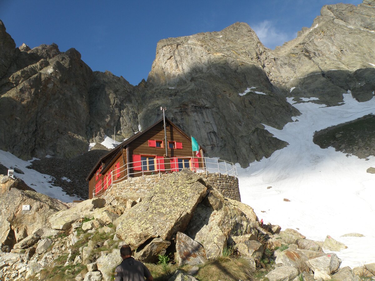 Fotografia: Il rifugio Bozano con struttura di legno e rosse imposte. Sullo sfondo la spettacolare parete sud-ovest del Corno Stella di forma trapezioidale.