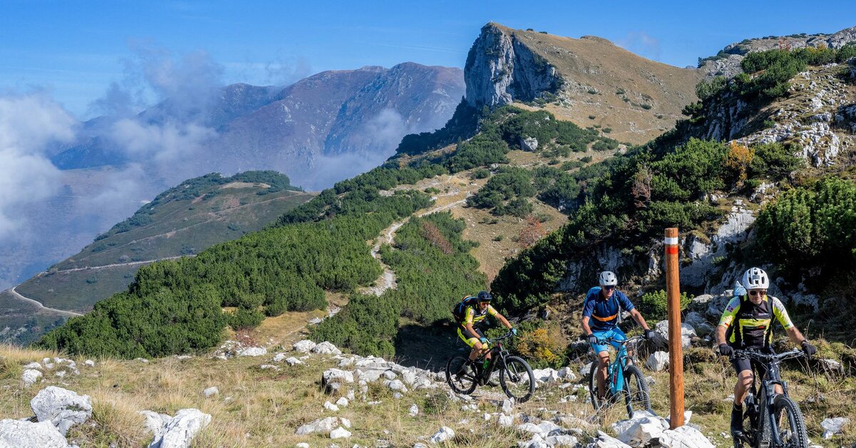 Fotografia: Tre ciclisti sulla ciclovia del Duca, nel Parco del Marguareis. Sullo sfondo si vede il passo del Duca e il sentiero di discesa che taglia a zig-zag il pendio erboso. Foto di Roberto Croci.