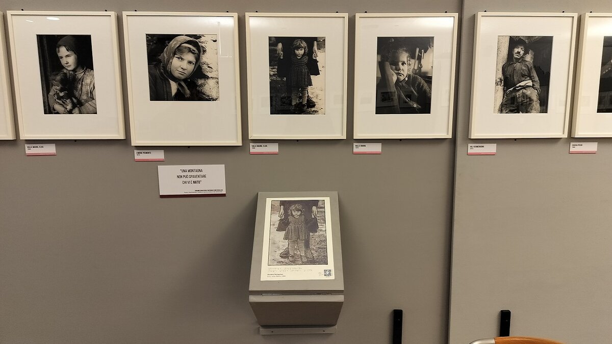 Sopra cinque scatti in bianco e nero di Michele Pellegrino, rappresentanti vari personaggi delle vallate del nostro territorio. Sotto, al centro, lo scatto tattile realizzato da CAMERA Torino, raffigurante una bambina con un vestito a quadri che tiene le mani a una donna, visibile solo in parte, che si trova dietro di lei. Lo scatto è caratterizzato dalla presenza di rilievi, da una descrizione in braille e da un QR Code in basso a destra che rimanda a un video descrittivo della fotografia.