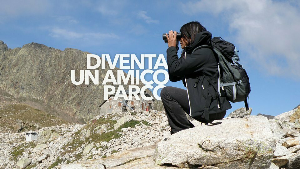 Nella fotografia: si ve una ragazza seduta che binocola davanti al rifugio Questa. In sovraimpressione la scritta Iscriviti a Amico Parco
