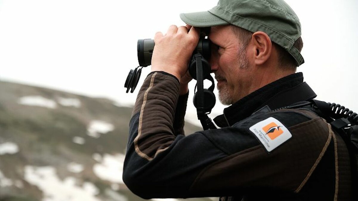 La fotografia mostra, in primo piano, un guardiaparco che tiene in mano un binocolo e guarda verso sinistra. Si nota il logo dell'Ente APAM sul braccio. Lo scatto è di I. Sigaudo