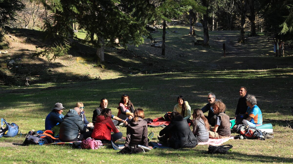 La foto mostra un gruppo di persone, in cerchio, sedute per terra sull'erba, attorno a loro un bosco. La foto è di Weco Impresa Sociale. Fine della descrizione.