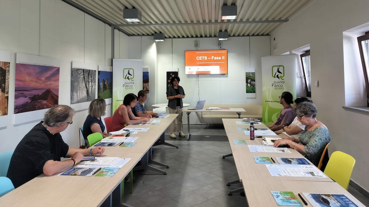 La fotografia mostra, sullo sfondo, uno schermo con sopra la presentazione della Fase 2 della CETS, alla sinistra Erika Chiecchio delle APAM intenta nell'esposizione del lavoro. Di fronte a lei, semigirati di spalle e seduti al tavolo, gli operatori che hanno partecipato all'incontro. Fine della descrizione.