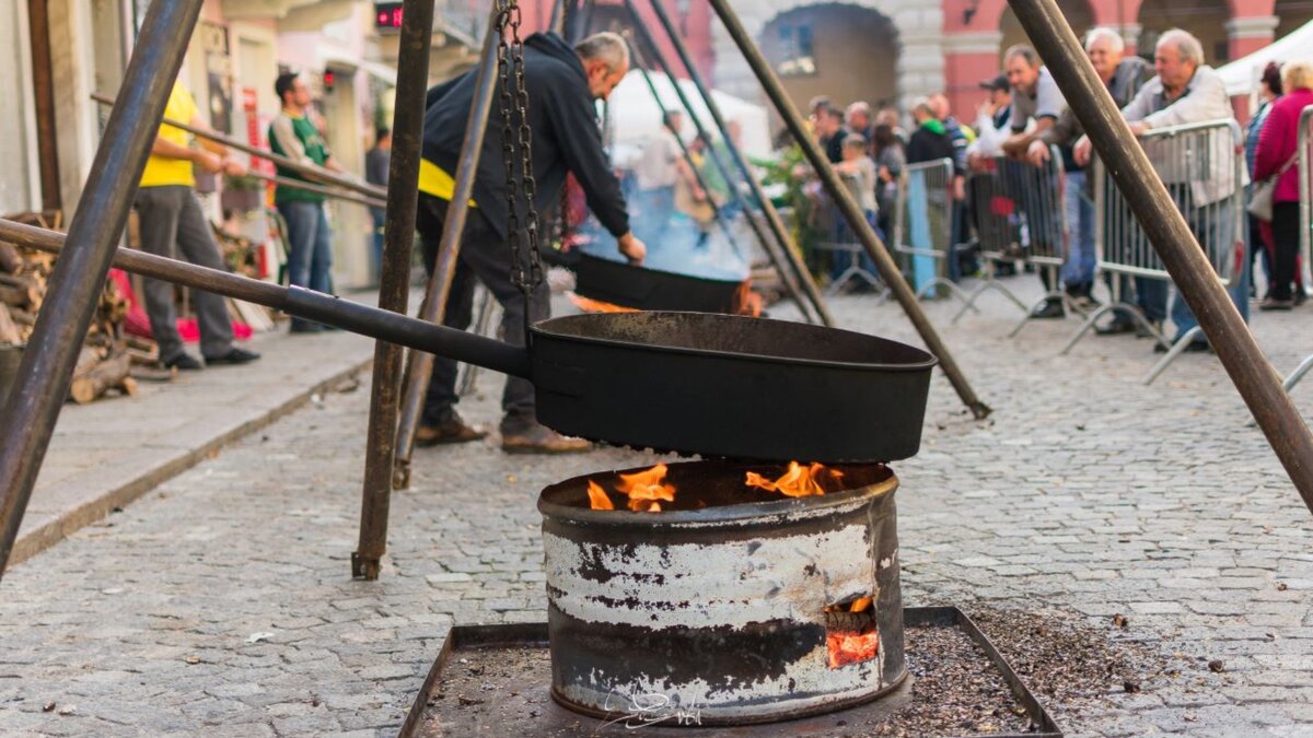 La foto mostra, in primo piano, la padella per fare le caldarroste, sullo sfondo un caldarrostaio che lavora. Fine della descrizione.