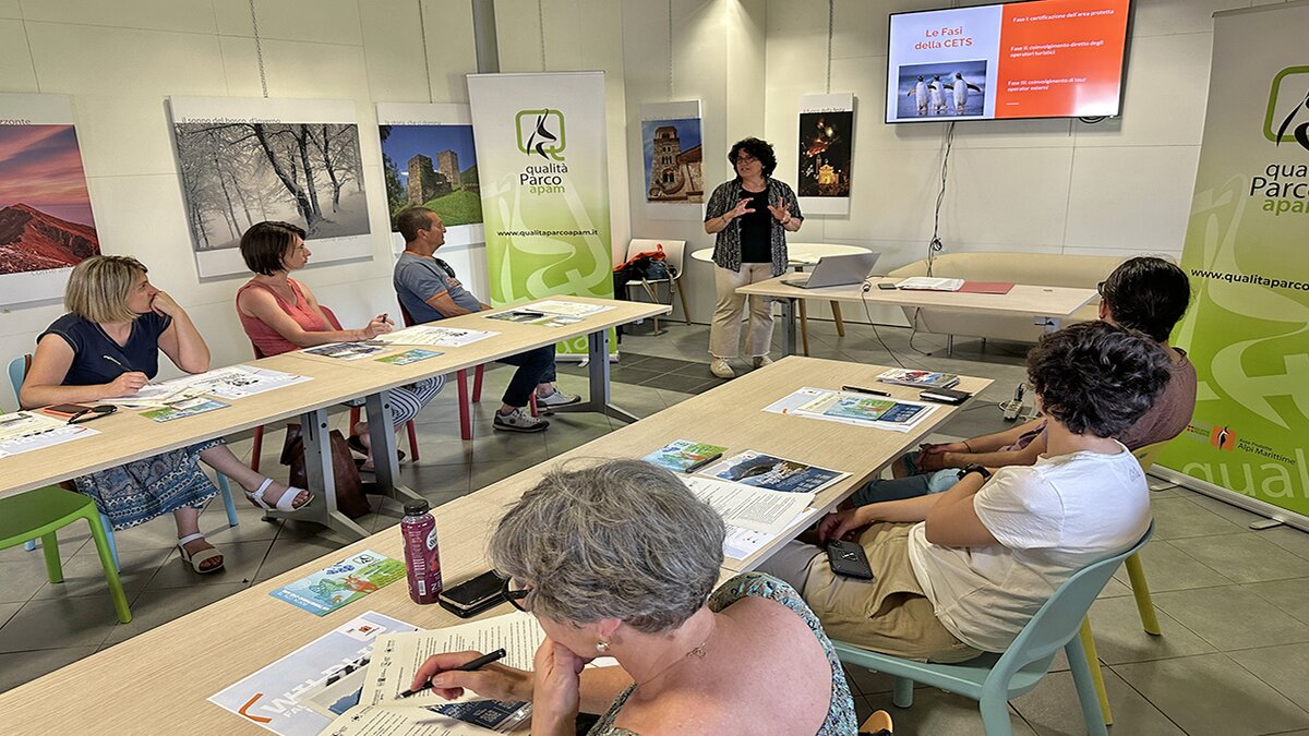 La foto mostra un gruppo di persone sedute a un tavolo che ascolta E. Chiecchio delle Aree Protette Alpi Marittime che illustra i principi della Fase II della CETS. Alle spalle della persona in piedi, le slide sulla CETS. Fine della descrizione.