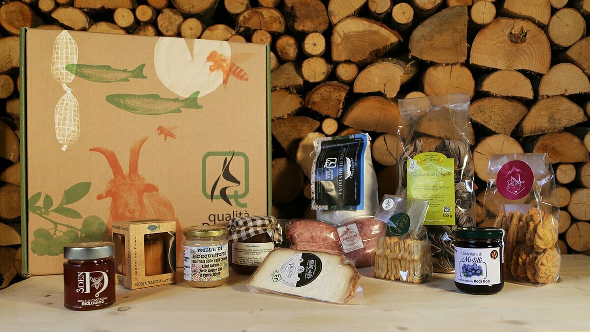 La foto mostra, sulla sinistra, la scatola che contiene i prodotti a marchio Qualità Parco APAM; di fronte, gli 11 prodotti contenuti nel Pacco Aquila reale, sullo sfondo una legnaia. Fine della descrizione.