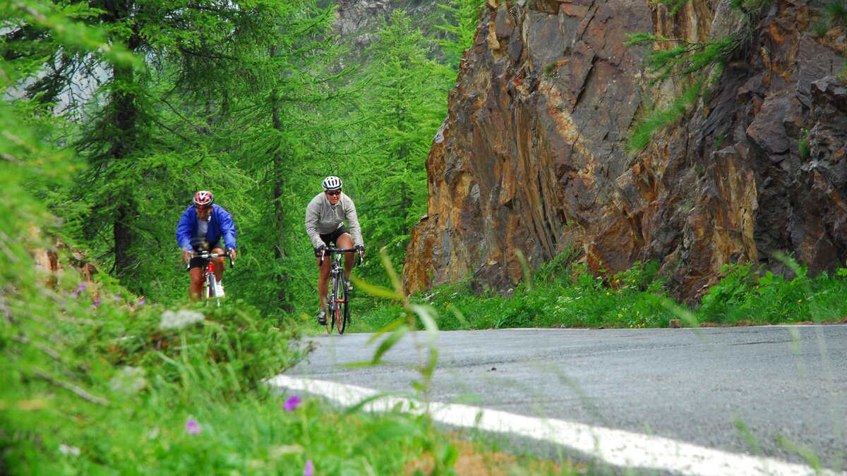 Descrizione foto Due ciclisti in salita sulle strade della Gran Fondo Margreen, La strada è bagnata e lo sfondo è una roccia rossastra | G. Bernardi Fine descrizione