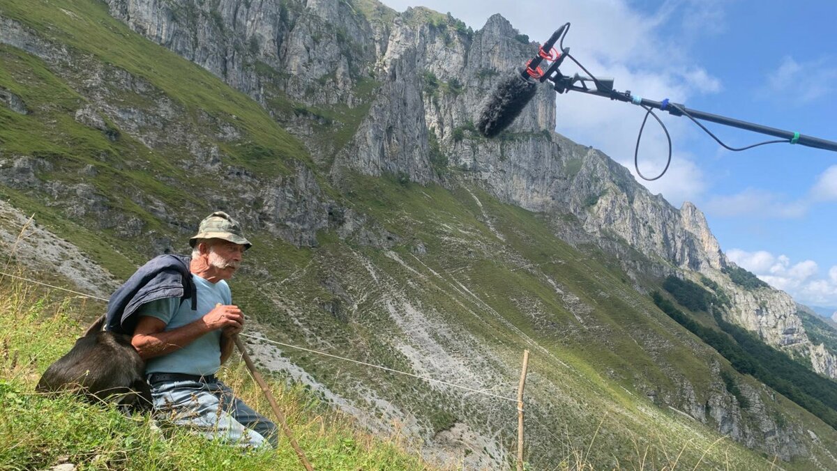 La foto mostra sulla sinistra un pastore seduto sull'erba, in alto a destra un microfono. Si tratta di uno scatto effettuato durante la registrazione di un filmato. Sullo sfondo le montagne.