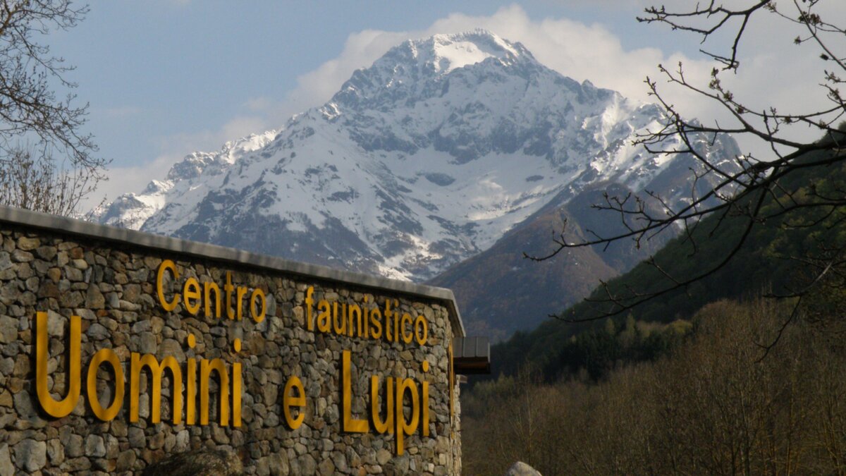 Lo scatto di A. Rivelli mostra, a sinistra, l'insegna dell'ingresso dell'Area faunistica Uomini e Lupi. A destra, sullo sfondo, le montagne innevate. Fine descrizione foto.
