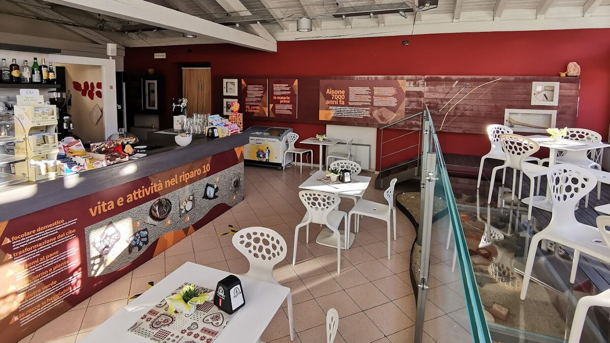 Nella foto si vede la sala del bar della Taverna delle Grotte con il bancone che con immagini e testo presentano il diorama che si trova davanti sotto un soppalco vetrato (su cui ci sono tavolini)| G. Bernardi Fine descrizione foto.