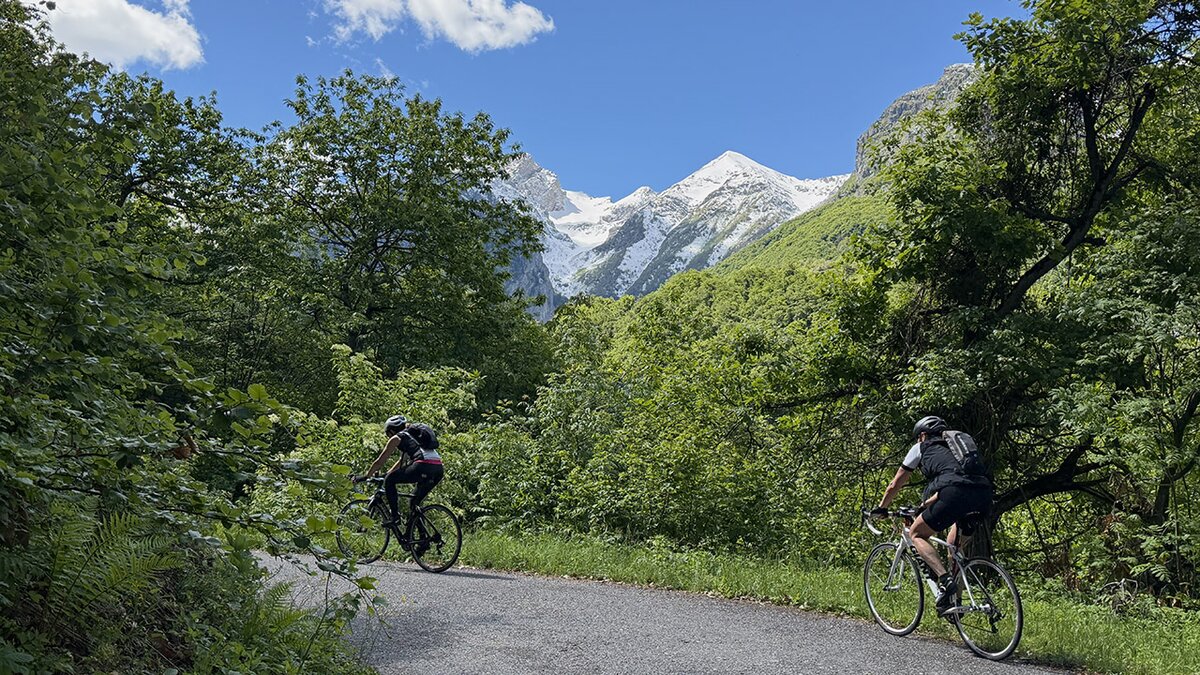 Descrizione foto: si vedono due cicliste in salita sulla strada immersa nel bosco con un'apertura che fa vedere il Monte Servantun tutto innevato. Fine descrizione foto.