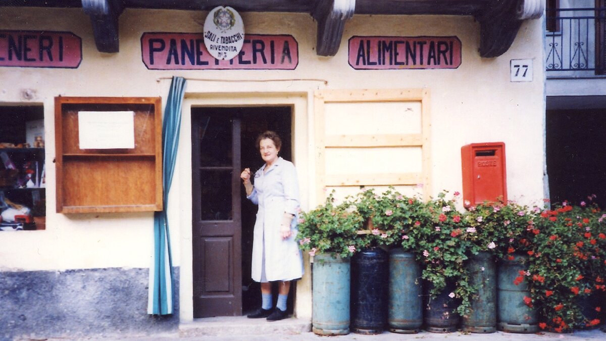 Marta sulla porta della //butéa//. Le lettere delle scritte panetteria e alimentari sono blu su fondo rosa. Alla sinistra di Marta, vasi di gerani sono sorretti da bombole del gas. Archivio famiglia Franco. Fine descrizione dell'immagine.